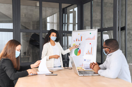 Diverse work team in protective medical masks working together in modern coworking space, multiracial employee brainstorming during pandemic period, a biracial woman presenting ideas. New social rules