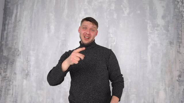 Young attractive male in a gray sweater speaks gesturing while standing against a gray wall. Thumbs up gesture, optimistic mood, people and emotions