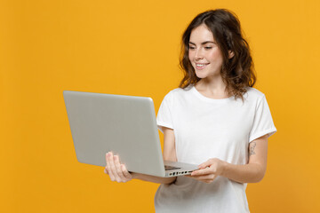 Naklejka premium Young smiling happy freelancer copywriter caucasian student woman in white basic t-shirt look camera hold laptop pc computer chat browsing isolated on yellow orange color background studio portrait.