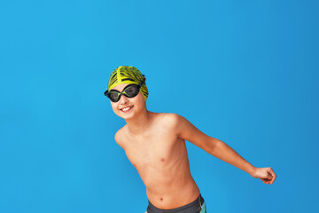 portrait of a sporty boy in a swimming cap, glasses and a bathing suit, performing exercises on a blue background. child enjoys swimming in a sports school. concept healthy lifestyle and sports.