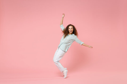 Full Length Young Black African American Happy Smiling Friendly Curly Woman In Blue Shirt Look Camera Standing On Toes Leaning Back Dancing Have Fun Isolated On Pastel Pink Background Studio Portrait.