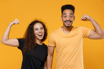 Young couple two american friends strong sporty fitness family african man woman in black t-shirt show biceps muscles on hand demonstrate strength power isolated on yellow background studio portrait.