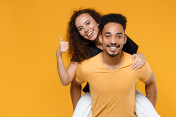 Young couple two together family smiling happy african man woman 20s in black t-shirt giving piggyback ride to joyful sit on back show thumb up gesture isolated on yellow background studio portrait.