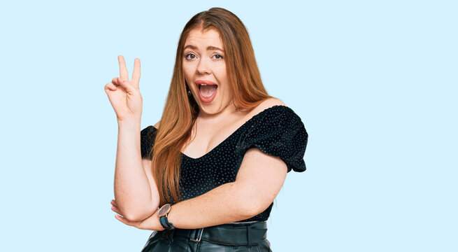 Young beautiful redhead woman wearing elegant clothes smiling with happy face winking at the camera doing victory sign. number two.