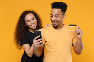 Young couple two friends together family african smiling happy man woman 20s in black t-shirt hold mobile cell phone credit bank card shop online isolated on yellow color background studio portrait