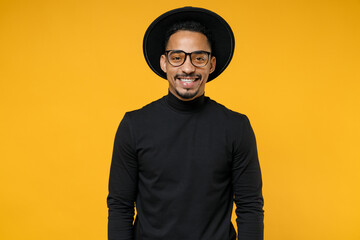 Young smiling attractive fashionable african american man 20s wearing stylish black hat shirt eyeglasses looking camera isolated on yellow orange background studio portrait. People lifestyle concept.