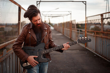 A stern man with a long red beard and gray hair in a brown leather jacket plays the black guitar and waves his head