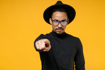 Young strict serious leader african american man 20s wear stylish black shirt hat eyeglasses point index finger camera on you command do it isolated on yellow orange color background studio portrait