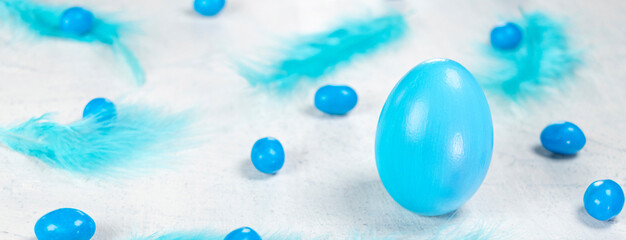 Banner with blue egg on a white concrete background with blue feathers and blue chocolates in the shape of an egg. Happy Easter concept.
