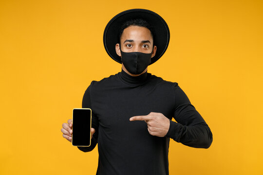 Young african man 20s in black hat shirt face mask from coronavirus covid-19 pandemic quarantine point finger on mobile phone with blank screen workspace isolated on yellow background studio portrait. - Powered by Adobe