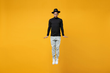 Full length of young active fashionable overjoyed fun happy excited fun african man in stylish black hat shirt eyeglasses jump high looking camera isolated on yellow color background studio portrait.