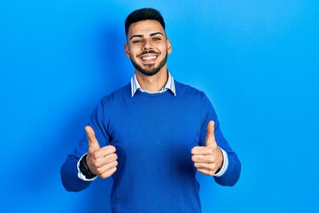 Young hispanic man with beard wearing casual blue sweater success sign doing positive gesture with hand, thumbs up smiling and happy. cheerful expression and winner gesture.