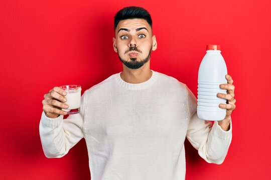 Young hispanic man with beard holding glass of milk and plastic bottle puffing cheeks with funny face. mouth inflated with air, catching air.