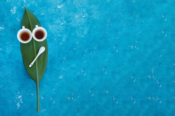 Two white cups with tea standing on a green leaf with blue background, concept face, view from top