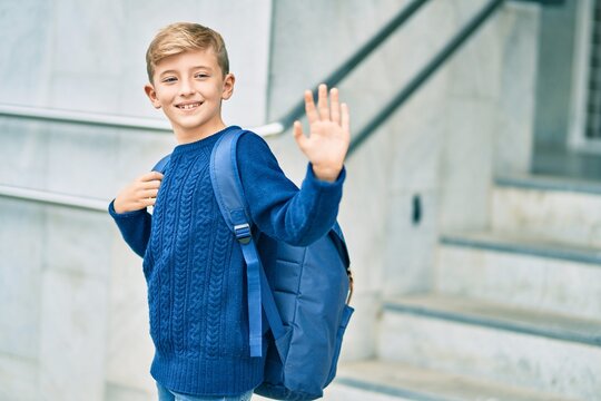 Adorable blond student kid saying goodbye with hand getting in the school.