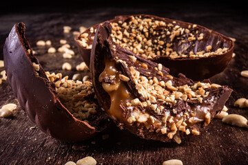 Stuffed chocolate easter egg with peanut and caramel on a wooden table with lots of peanuts around. Cracked. Close up.