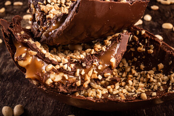 Stuffed chocolate easter egg with peanut and caramel on a wooden table with lots of peanuts around. Cracked. Close up.