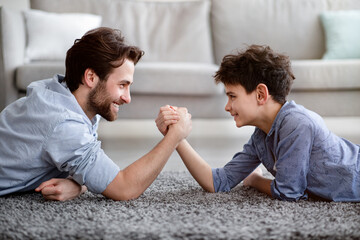 Happy father competing in arm-wrestling with his son, enjoying time together at home, side view - obrazy, fototapety, plakaty
