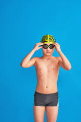 small swimmer wearing swimming cap, glasses and bathing suit on blue background. child adjusts his glasses, water go. boy is studying at sports school, learning to swim. Problems with swimming goggles