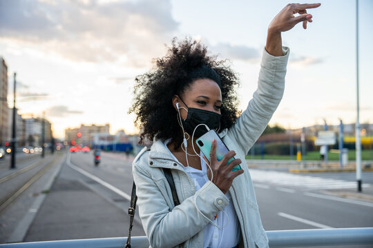 Portrait of a young woman in the evening at sunset in the city wearing protective face mask during global pandemic from Covid-19 Coronavirus dancing listening to music with earphones from smartphone