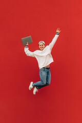 Vertical photo of a guy in stylish casual clothes isolated in a jump on a red background, looking at the camera with a smile on his face. The guy with a laptop is levitating on a red background.