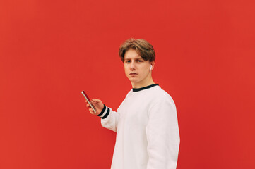 Handsome young man in wireless headphones with a smartphone in his hand stands on a background of a red wall and looks at the camera with a serious face.