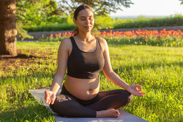 Cute young caucasian pregnant woman is meditating while sitting on a rug on the lawn on a sunny...