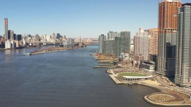 A high angle view looking north over the East River on a sunny day. The drone camera dolly out and pan left, looking up towards the Upper East side of Manhattan, NY.