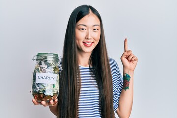 Obraz premium Young chinese woman holding charity jar with money smiling with an idea or question pointing finger with happy face, number one