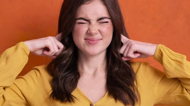 A displeased young woman is covering her ears with her fingers standing isolated over orange background in the studio