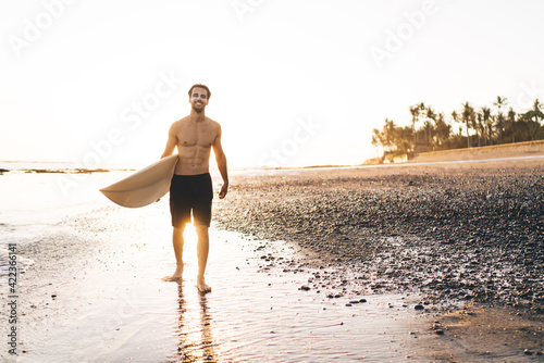 Full Length Of Man With Sexy Body Holding Surfboard Ready For Amateur Rides  In Ocean, Happy Caucasian Male Surfer Smiling At Camera Posing At Coastline  While Recreating At Kuta, Bali Indonesia Canvas