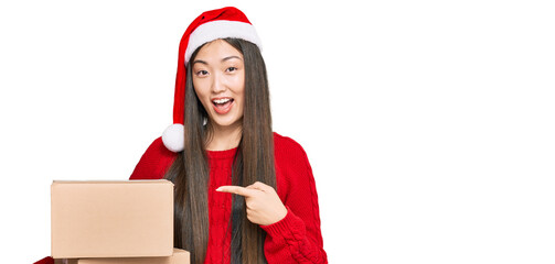 Young chinese woman wearing christmas hat holding delivery package smiling happy pointing with hand and finger