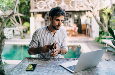 Focused man with notebook and laptop sitting near swimming pool
