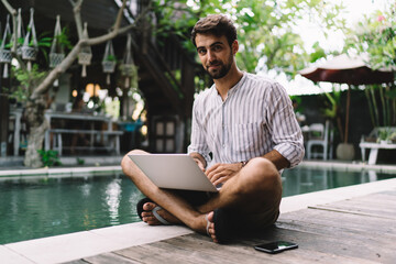 Positive young man working distantly on laptop during summer vacation in tropical resort