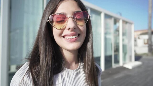 Happy beautiful young teenager with long hair, and plump lips wearing funny heart-shaped sunglasses laughing looking at camera in city outdoor commercial centre. Concept of confidence in the future