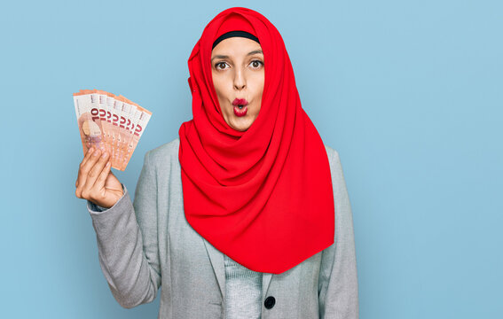 Beautiful hispanic woman wearing islamic hijab holding 10 euros banknotes scared and amazed with open mouth for surprise, disbelief face