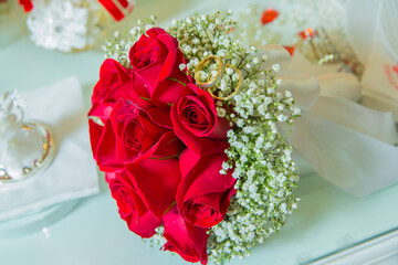 Red ribbon . Red engagement flowers on white table . Bride and groom with Engagement gold rings put on the red wedding bouquet . Red engagement bouquet .