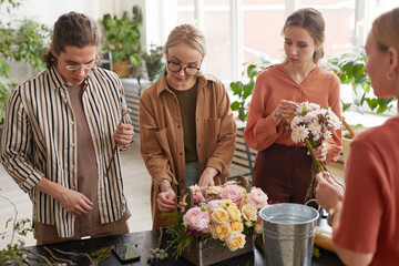 Diverse group of young people arranging flowers in florists workshop while attending class on floral art