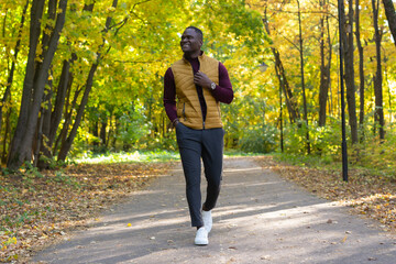 Fototapeta premium Cheerful african american young man in stylish clothes walks in autumn park on sunny warm autumn day. Concept of weekend and outdoor recreation