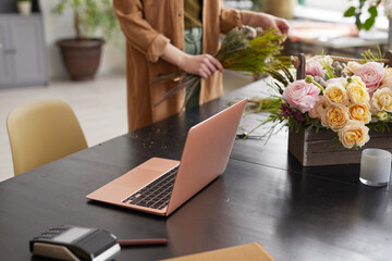Close up of open laptop on table in flower shop with unrecognizable female florist in background,...