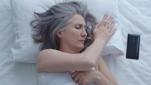 Mature sleepy lady looking at time on smartphone, hiding under blanket, laziness