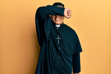 Young latin priest man standing over yellow background covering eyes with arm smiling cheerful and funny. blind concept.