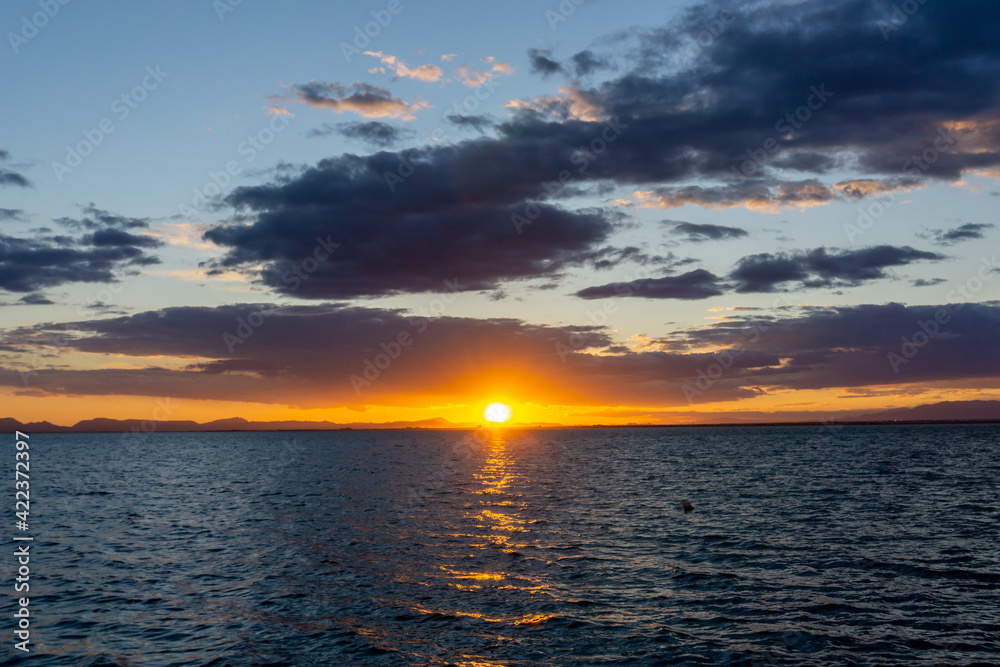 Sticker scenic and colorful sunset over the ocean with shimmering light on the waves and silhouette of mountains on the shore behind