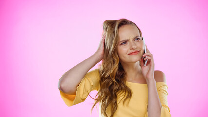 displeased young woman adjusting wavy hair while talking on smartphone on pink