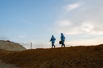 Figures of two contemporary ecologists in protective coveralls moving on top of hill with dirty soil against cloudy sky during investigation