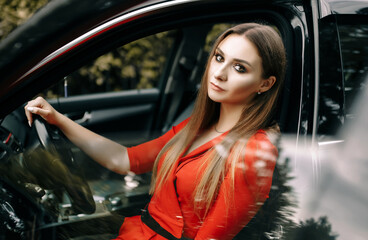Naklejka na ściany i meble A beautiful young girl in a red overalls sits behind the wheel of a black car on an empty road in the forest