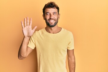 Handsome man with beard wearing casual yellow tshirt over yellow background showing and pointing up with fingers number five while smiling confident and happy.