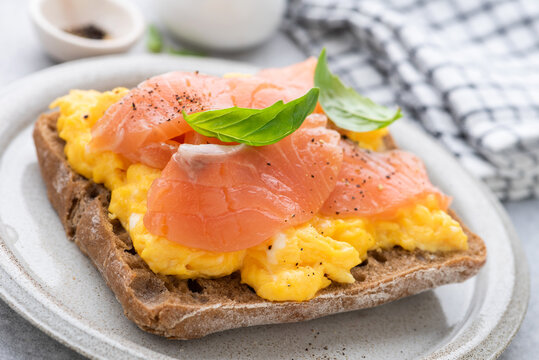 Rye bread toast with scrambled eggs and smoked salmon garnished with black pepper and basil leaf. Tasty breakfast open sandwich with egg and lox
