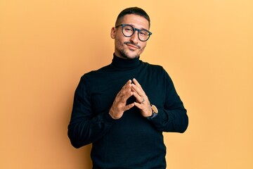 Handsome man with tattoos wearing turtleneck sweater and glasses hands together and fingers crossed...