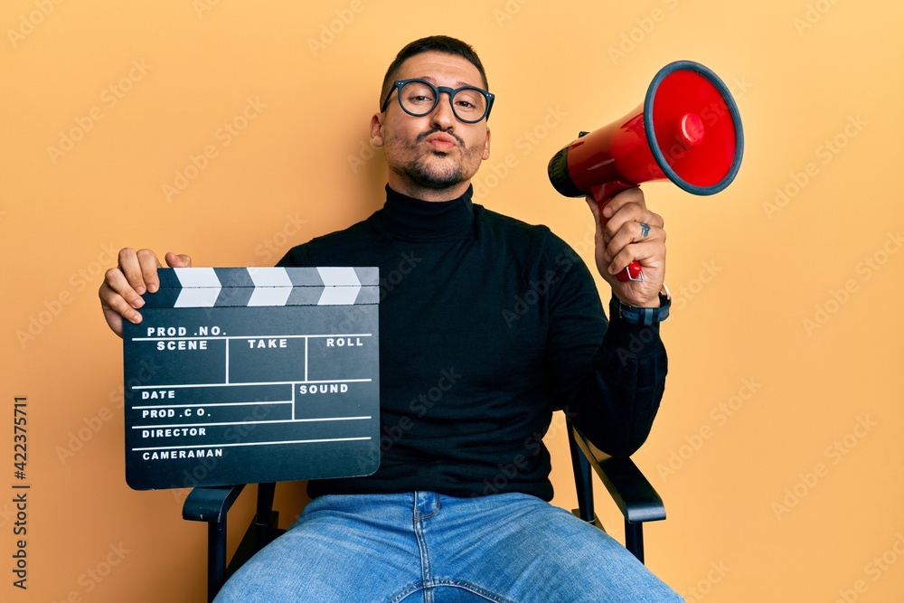 Poster handsome man with tattoos holding video film clapboard and megaphone looking at the camera blowing a